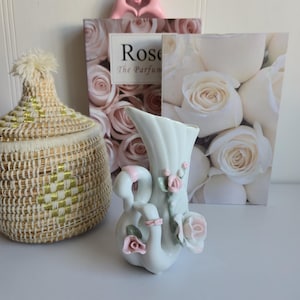 May include: A white ceramic vase with swan-shaped handles and pink rose accents. A woven basket with a gold and beige pattern and a book titled "Rose The Parfum" are also in the image. The background features a white wall.
