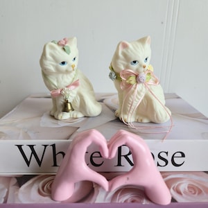 Peut inclure: Deux figurines de chat en céramique crème avec des yeux bleus et des détails floraux. Un chat a une cloche sur son collier, l'autre un nœud rose. Un cœur rose en forme de main est devant les chats, avec le texte "White Rose" et "Pink Rose" visible.