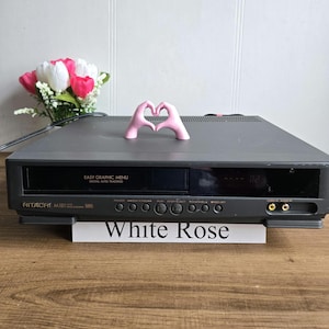 Puede incluir: Un reproductor de VHS Hitachi M181 gris con el texto "EASY GRAPHIC MENU" y "White Rose". Una escultura de mano rosa en forma de corazón está encima. Un ramo de flores rosas y blancas está en el fondo.