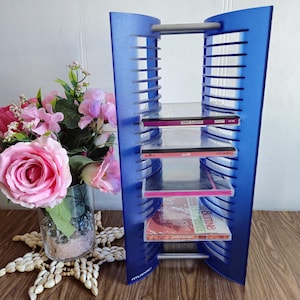 May include: A blue plastic CD storage tower with a silver metal handle. The tower has four shelves and is holding several CDs. The tower is sitting on a wooden surface.