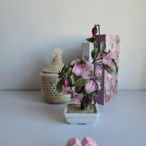 Peut inclure: Un petit bonsaï avec des fleurs roses et des feuilles vertes dans un pot carré blanc. Un panier en osier et un livre avec un motif de roses sont en arrière-plan. Une sculpture rose en forme de cœur est au premier plan.