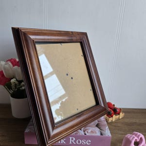 Peut inclure: Un cadre photo rectangulaire en bois marron foncé. Le cadre est vide, avec un dos en carton. Le cadre repose sur une boîte rose avec le texte "K Rose".