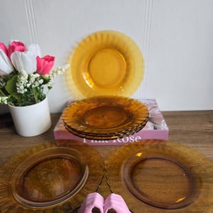 Puede incluir: Platos de vidrio color ámbar con borde festoneado. Los platos están dispuestos sobre una superficie de madera, con una pila de platos en el fondo. Un adorno rosa en forma de mano está en primer plano.