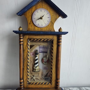 Puede incluir: Un reloj de pared de madera con temática náutica. La esfera del reloj está rodeada por un marco de madera con un borde azul. La esfera del reloj tiene números romanos y una aguja negra. El reloj está montado sobre una base de madera con un borde azul. La base tiene una ventana de vidrio que muestra un faro en miniatura, un salvavidas y una red de pesca. El reloj es una gran adición a cualquier hogar con temática náutica.