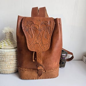 Könnte beinhalten: Brauner Lederrucksack mit geprägtem Palmenmuster auf der Klappe. Die Tasche hat einen Kordelzugverschluss, einen geflochtenen Boden und einen Schnallenverschluss. Der Rucksack ist etwa 38 cm hoch.