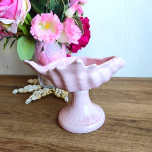 Peut inclure: Un bol en céramique rose en forme de coquille de mer avec un piédestal. Le bol est posé sur une surface en bois.