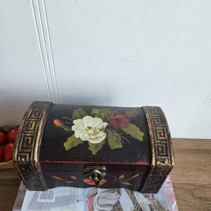 May include: A small, dark wooden chest with a curved top and decorative gold trim. The chest features a hand-painted floral design in white, red, and green. It has a metal clasp and is resting on a stack of books.