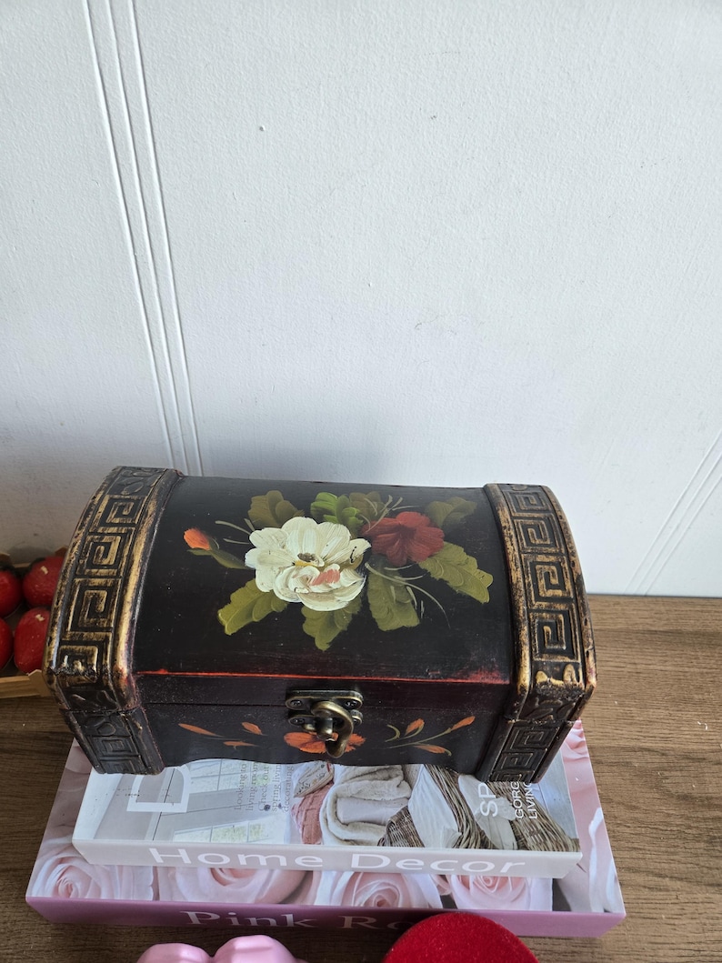 May include: A small, dark wooden chest with a curved top and decorative gold trim. The chest features a hand-painted floral design in white, red, and green. It has a metal clasp and is resting on a stack of books.