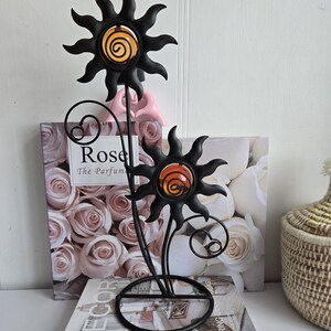 Könnte beinhalten: Schwarzer Metall-Sonnen-Kerzenhalter mit orangefarbenen und gelben Glaszentren. Das dekorative Stück hat ein florales Design mit kreisförmiger Basis und einem Wirbelakzent. Der Hintergrund enthält ein Buch mit dem Titel "Home Decor".