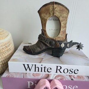 Puede incluir: Un marco de fotos decorativo con forma de bota de vaquero con espuela. El marco es marrón y beige con un aspecto de cuero envejecido. La abertura del marco es de 6,35 cm x 8,89 cm. El marco está sobre una pila de libros con el texto "White Rose".