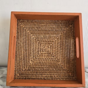 Op de afbeelding: Een vierkant geweven dienblad met een natuurlijke rieten basis en een helder oranje houten frame. Het dienblad heeft twee kleine handvatten aan de zijkanten.