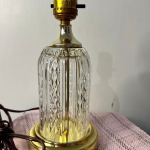 Puede incluir: Una lámpara de mesa de cristal transparente con una base dorada y un patrón de diamante. La lámpara tiene un cable y un casquillo de bombilla estándar.