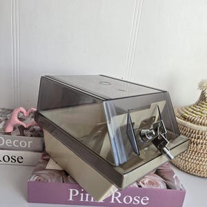 Peut inclure: Une boîte en plastique beige et grise avec un couvercle transparent incliné. La boîte a une serrure avec une clé insérée. La boîte est posée sur un livre avec les mots "Pin Rose" sur la couverture.