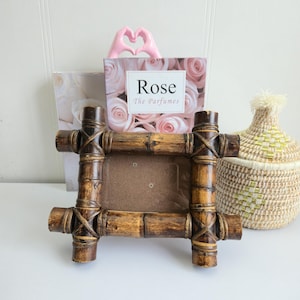 Peut inclure: Un cadre photo en bambou avec une ouverture rectangulaire, finition marron naturel. Une boîte avec le mot "Rose" et un geste de la main en forme de cœur sont derrière le cadre. Un panier tressé est à droite.
