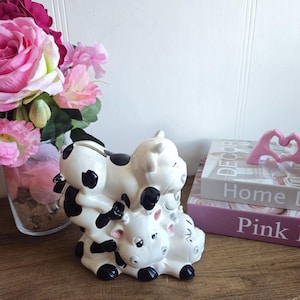 May include: A ceramic money box shaped like three stacked cows. The cows are black and white with pink noses and black eyes. The top cow is lying down with its head resting on the middle cow's back.
