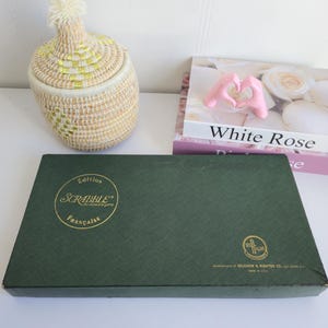 Pode incluir: Uma caixa verde do jogo de tabuleiro Scrabble com letras douradas e uma cesta tecida com tampa. A caixa é retangular e tem as palavras "Edition Scrabble Française" impressas. Um livro com o título "White Rose" também é visível.
