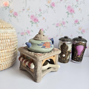 May include: A ceramic oil warmer featuring a light green teapot-shaped top with a painted orange flower, resting on a square beige base. Beside it are two cylindrical candles in ornate black and gold holders with small white pearls.