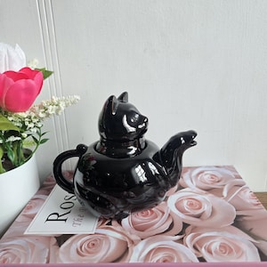 May include: A black ceramic cat-shaped teapot. The teapot has a rounded body, a curved spout, and a handle. The cat's head forms the lid, with ears and a face. The teapot is placed on a book titled "Pink Rose".