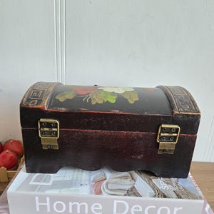May include: A small, dark brown wooden chest with a curved top and brass-colored clasps. The top is painted with a floral design in red, white, and green. The chest is sitting on a stack of books, one of which reads "Home Decor".