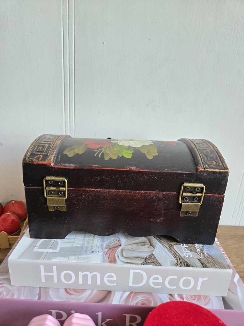 May include: A small, dark brown wooden chest with a curved top and brass-colored clasps. The top is painted with a floral design in red, white, and green. The chest is sitting on a stack of books, one of which reads "Home Decor".