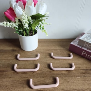 Op de afbeelding: Vijf roze plastic ladegrepen met een afgeronde rechthoekige vorm. De grepen zijn in een verspringend patroon op een houten oppervlak gerangschikt.