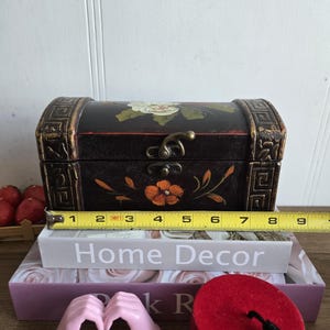 May include: A small, black wooden chest with floral designs and gold accents, measuring approximately 8 inches long. The chest is placed on top of decorative books, including one that reads "Home Decor."