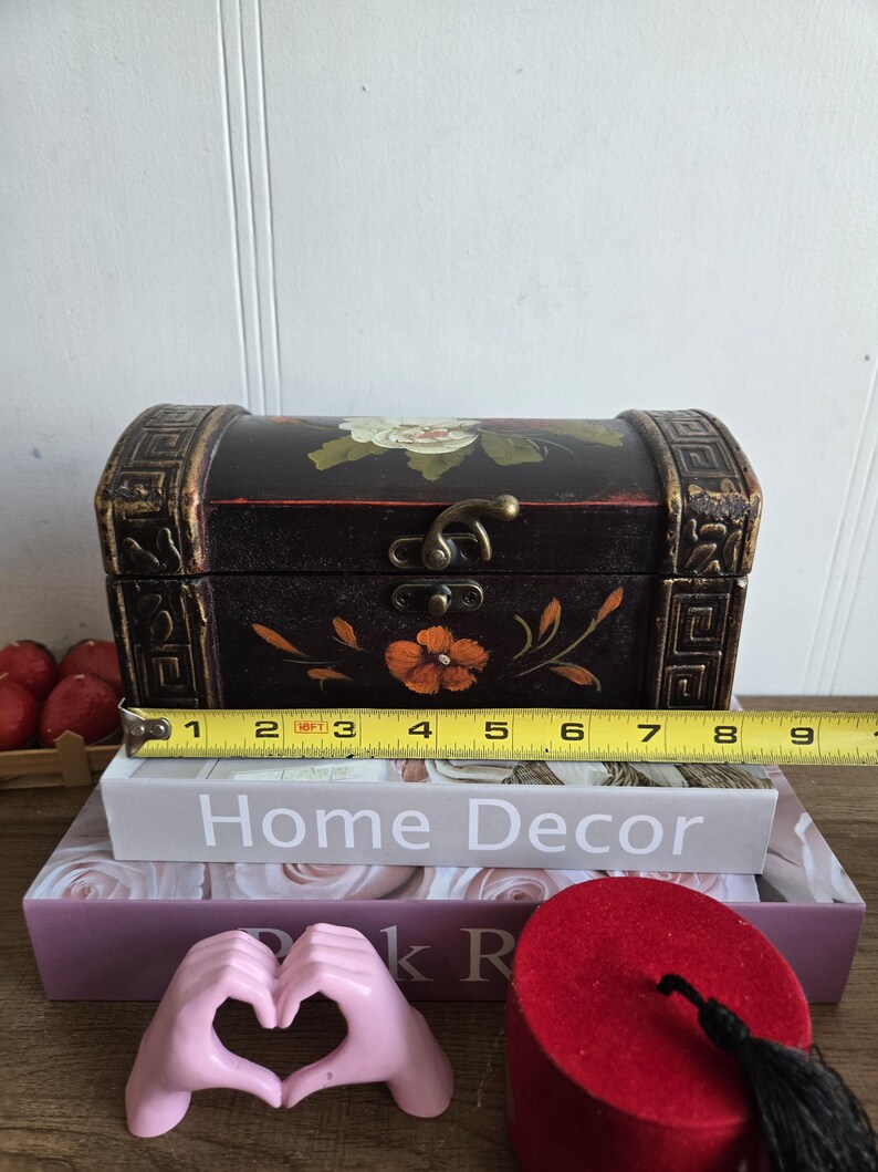 May include: A small, black wooden chest with floral designs and gold accents, measuring approximately 8 inches long. The chest is placed on top of decorative books, including one that reads "Home Decor."