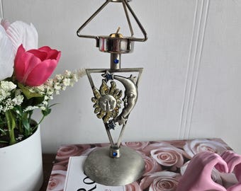May include: A silver-toned candle holder with a celestial design. The holder features a sun and moon motif, with a small candle at the top. The base is round, and the design includes blue accents. The candle holder is on a book with a floral design.