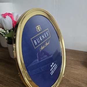 Peut inclure: Cadre photo ovale en laiton doré avec intérieur bleu foncé. Le cadre porte l'inscription "BURNES State Street" et mesure 20,3 x 25,4 cm. Le cadre est recouvert d'un vernis résistant au ternissement.