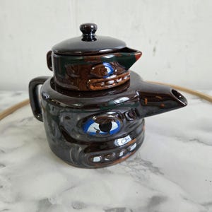 Puede incluir: Una tetera de cerámica marrón con una tapa en forma de cara con ojos azules. La tetera tiene un pico y una asa.