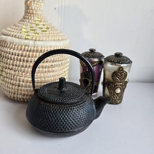 Peut inclure: Une théière en fonte noire avec une surface texturée et une poignée incurvée. À côté, deux bougeoirs décoratifs et un panier tressé avec un couvercle. Les objets sont disposés sur une surface blanche.