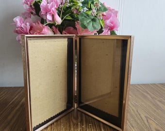 May include: A gold-toned double picture frame with two empty openings. The frame is made of wood and has a simple design. The frame is sitting on a wooden surface.