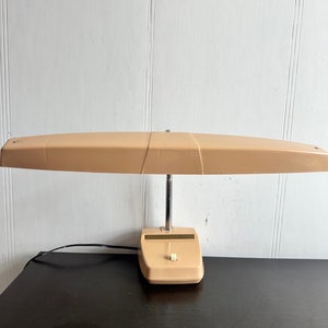 May include: A vintage beige desk lamp with a long, rectangular shade and a round base. The lamp has a chrome arm and a single switch on the base.