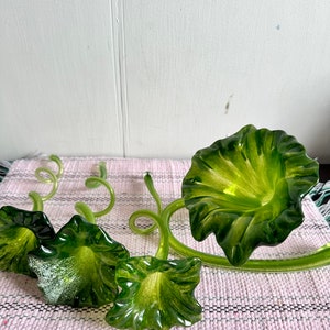 May include: A set of three green glass floral sculptures with curled stems and trumpet-shaped flowers. The sculptures are arranged on a pink and white striped surface.