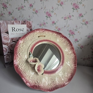 Peut inclure: Un miroir mural rond en céramique avec un centre crème et une bordure festonnée teintée de rose ornée de motifs floraux en relief. Deux fleurs sculptées en trois dimensions, blanches et roses, sont fixées sur le côté inférieur gauche.