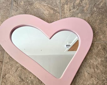 Puede incluir: Un espejo rosa en forma de corazón con un borde biselado. El espejo refleja un techo blanco y una pared blanca.