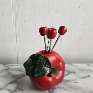 Puede incluir: Porta palillos de dientes en forma de manzana roja con cuatro palillos. La manzana tiene una hoja verde y está hecha de cerámica.