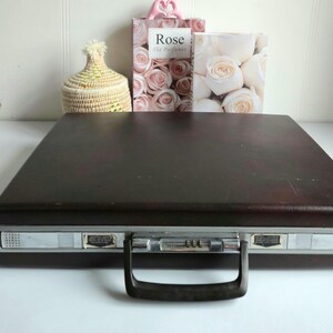 Puede incluir: Un maletín Samsonite vintage marrón oscuro con ribetes plateados y un asa negra. El maletín tiene una cerradura de combinación y está cerrado. En el fondo hay dos cajas con motivos florales y una cesta tejida.
