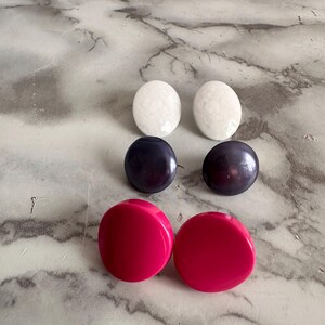 Peut inclure: Un ensemble de six boucles d'oreilles rondes en trois couleurs : blanc, noir et rose. Les boucles d'oreilles sont en plastique et ont une finition lisse et brillante.