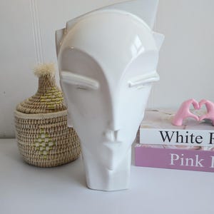 May include: A white ceramic face sculpture with a stylized design. The sculpture has a smooth, glossy finish and features closed eyes and a defined nose and mouth. A woven basket and books are in the background.