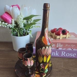 May include: A hand-painted wooden bottle and shot glass set. The brown bottle and glasses are decorated with pink, yellow, and orange floral designs. The set includes a round tray and is displayed with artificial flowers.