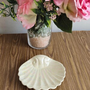 Peut inclure: Un plat en céramique blanche en forme de coquille de mer avec une finition nacrée. Le plat est posé sur une surface en bois.