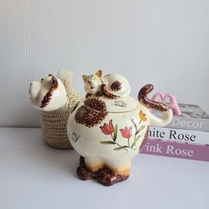 Peut inclure: Un pot à biscuits en céramique fantaisiste en forme de chat, avec un couvercle assorti. Le pot est de couleur crème avec des taches marron et des motifs floraux. Un récipient en forme de chien et des livres sont en arrière-plan.