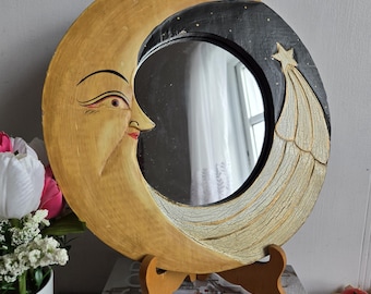 May include: A round, hand-painted wooden mirror in the shape of a crescent moon. The moon has a smiling face with a gold and black color scheme. The mirror is supported by a wooden stand.