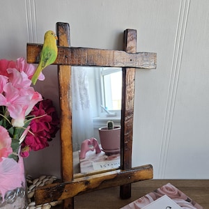 May include: A rustic wooden mirror with a distressed brown finish. The mirror is framed with thick wooden planks and has a small yellow bird perched on top. The mirror is sitting on a table with a box of rose-scented perfume.