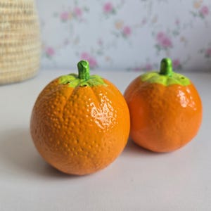 Peut inclure: Deux salières en céramique de forme ronde, colorées en orange et ressemblant à des agrumes. Chaque salière présente une surface texturée et alvéolée avec une petite tige vert vif sur le dessus. Elles ont une finition brillante.