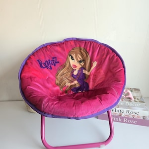 Peut inclure: Chaise pliante Bratz rose et violette. Le siège rond est orné d'un personnage Bratz et du logo. Le cadre est de couleur rose vif.