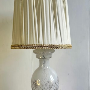 May include: White ceramic table lamp with a pleated white lampshade trimmed with gold. The lamp base has a delicate floral pattern.