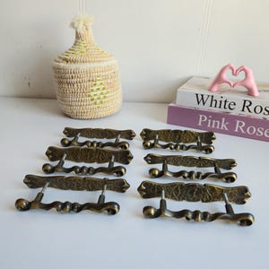 Peut inclure: Huit poignées de tiroir antiques en laiton sont présentées. Chaque poignée comporte une plaque rectangulaire texturée avec des détails décoratifs, reliée à une poignée incurvée. Un panier tissé avec un pompon et deux livres avec les titres "White Ros" et "Pink Ros" sont également dans l'image.