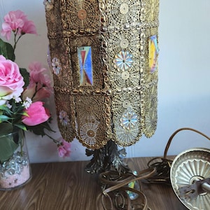 Può includere: Un paralume per lampada in metallo dorato con un design decorativo e intricato. Il paralume è decorato con accenti in vetro iridescente e presenta un motivo floreale. Il paralume è fissato a una base in metallo dorato con un motivo floreale.